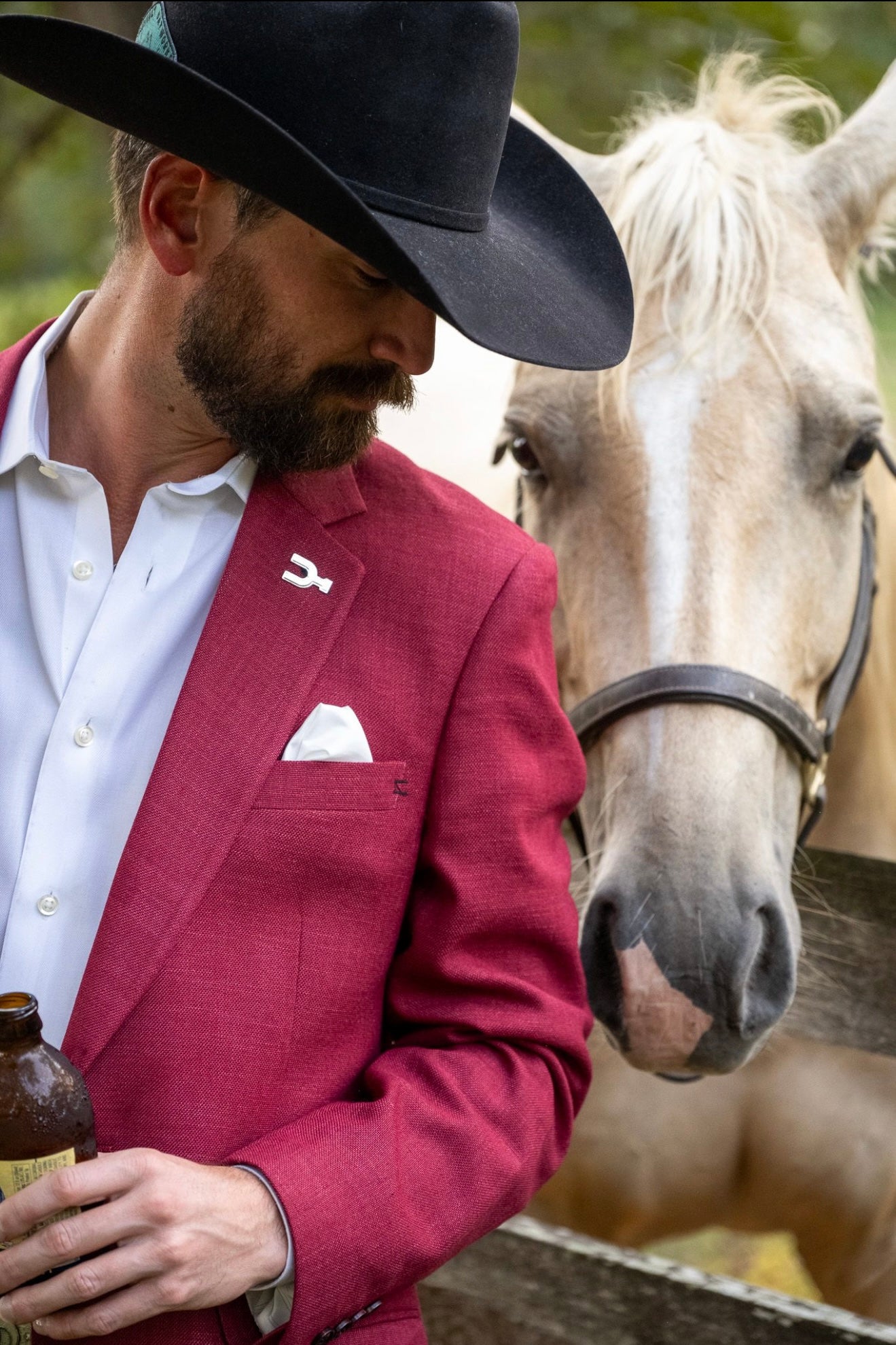Man in a red blazer and black cowboy hat standing next to a horse.