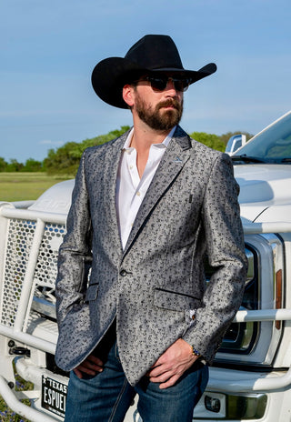 Man wearing a patterned blazer, white shirt, and black cowboy hat in front of a white truck.