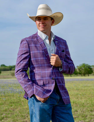 Man wearing a purple plaid jacket, white shirt, and cowboy hat standing in an open field.