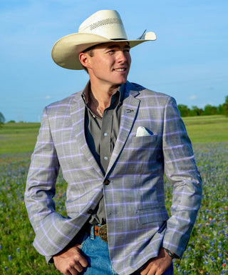 Man wearing a plaid grey and purple blazer, cowboy hat, and jeans standing in a field with blue sky and blue bonnets. Texas cowboy showcasing the new summer collection of western sport coats. Perfect for weddings or cowboy functions. 