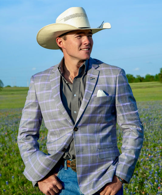 Man wearing a plaid grey and purple blazer, cowboy hat, and jeans standing in a field with blue sky and blue bonnets. Texas cowboy showcasing the new summer collection of western sport coats. Perfect for weddings or cowboy functions. 