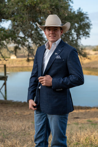 Man in a cowboy hat and blue suit standing by a body of water with trees in the background
