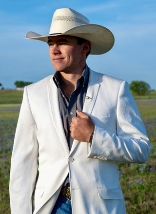 Man wearing a white blazer, blue shirt, and cowboy hat in an outdoor setting.