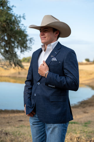 Man wearing a navy blazer, white shirt, and cowboy hat by a body of water.